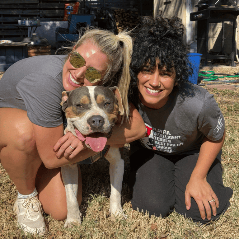 Two women kneeling and hugging a pitbull dog.