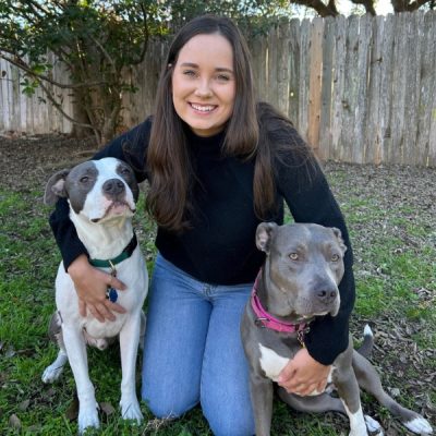 Woman kneeling in the grass with two pitbull dogs in front of her.