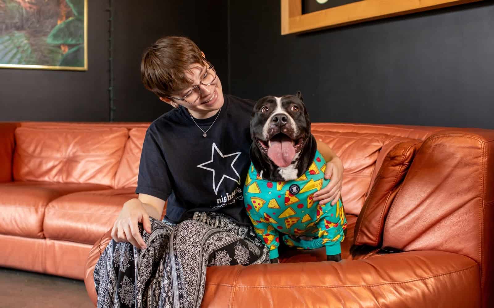 A woman sitting on a leather couch with a black pit bull dog wearing pajamas